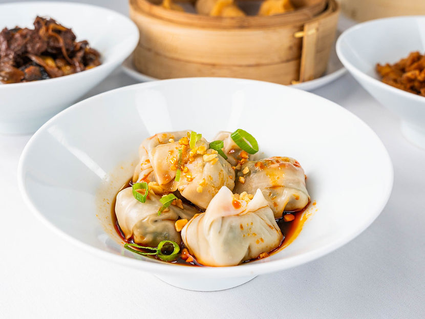 Steamed dumplings in white bowl with sauce, other dishes in the background.